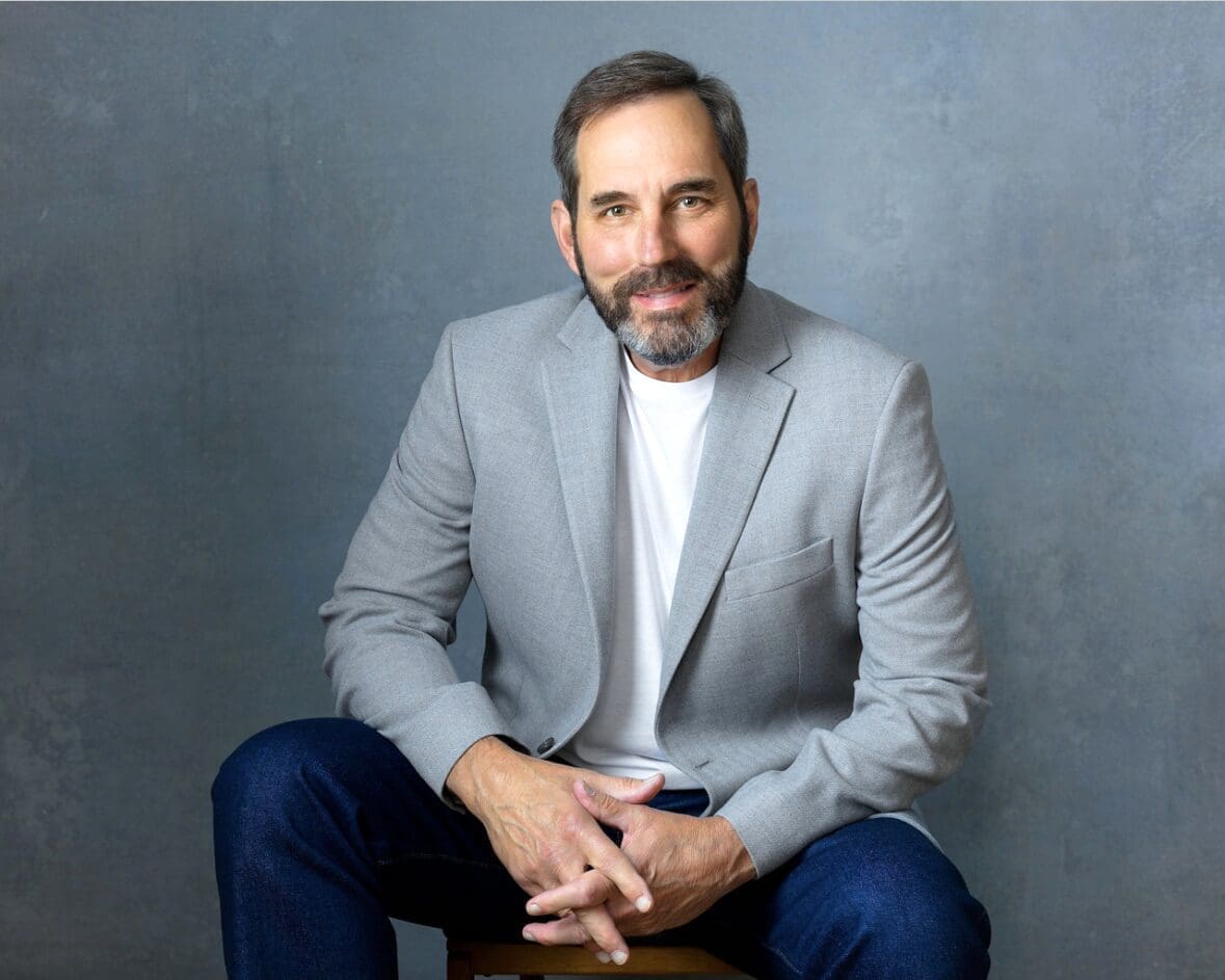 Man in a gray blazer and white shirt sitting against a gray background.
